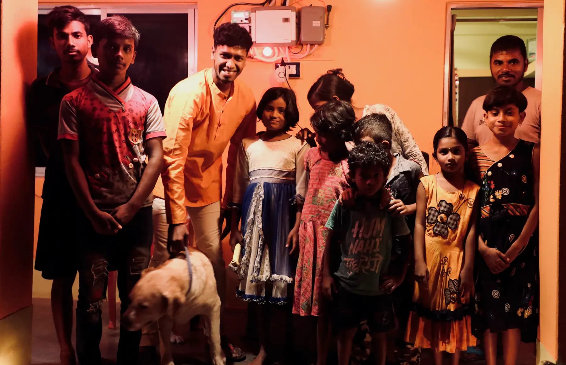 Village children and young men with a dog, gathered at night outside an orange-lit house at Turiyo
