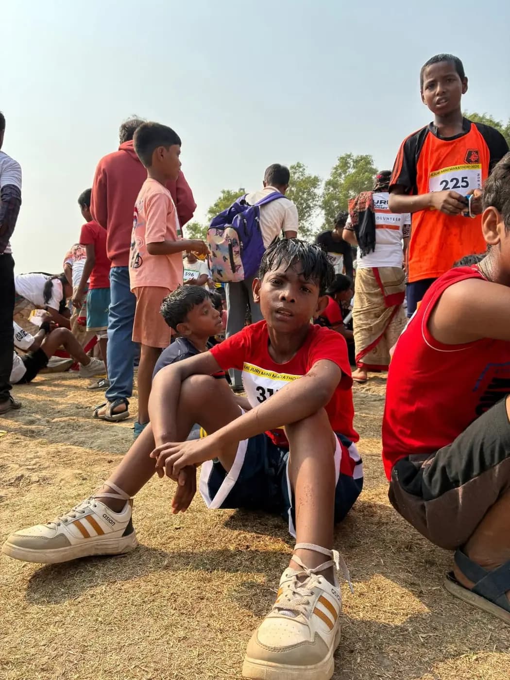 A boy in a red T-shirt and race bib sits on the dry ground catching his breath after the run, other runners around him