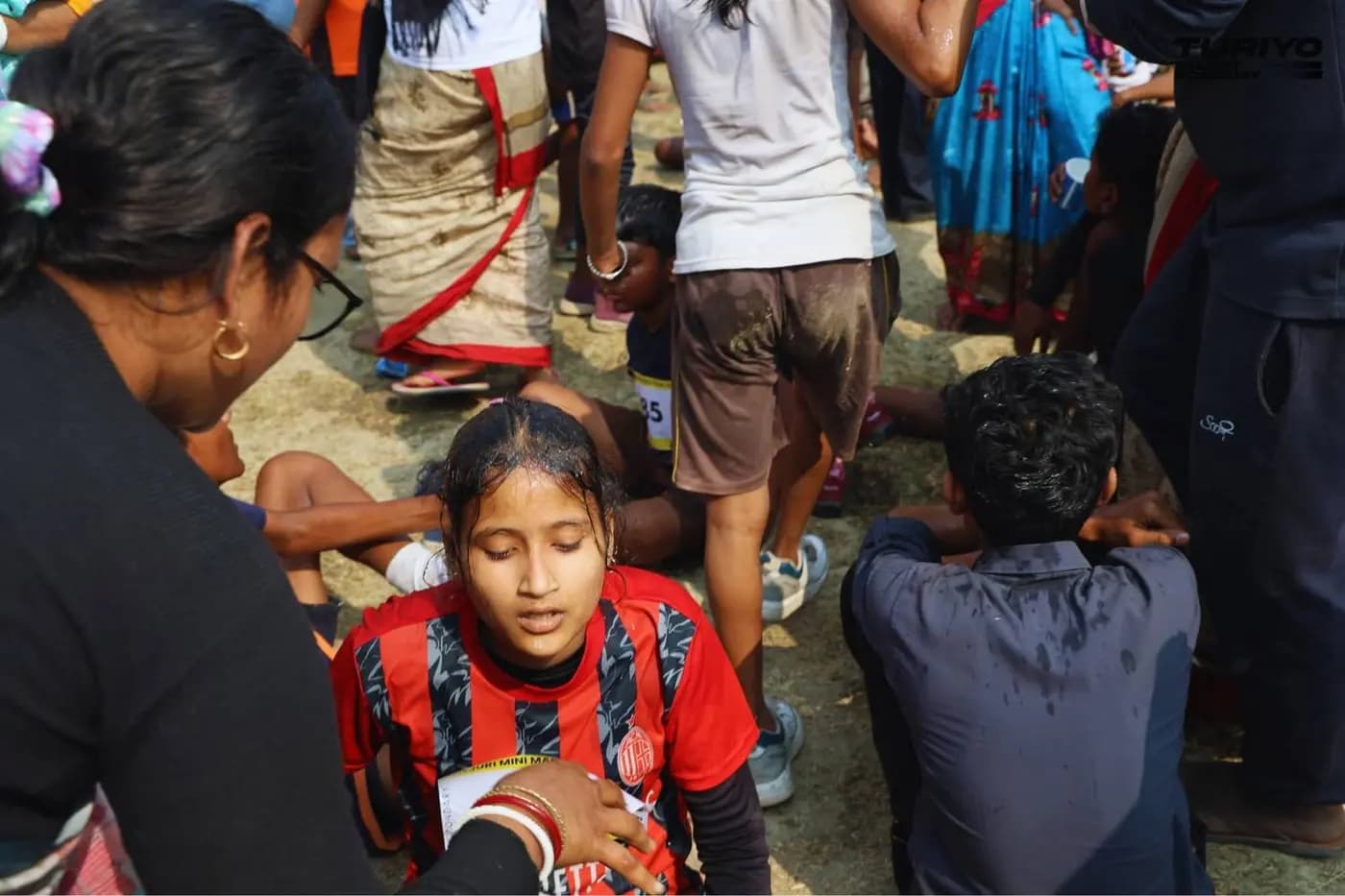 A woman looks after a girl runner sitting on the ground, hair soaked, just after the finish