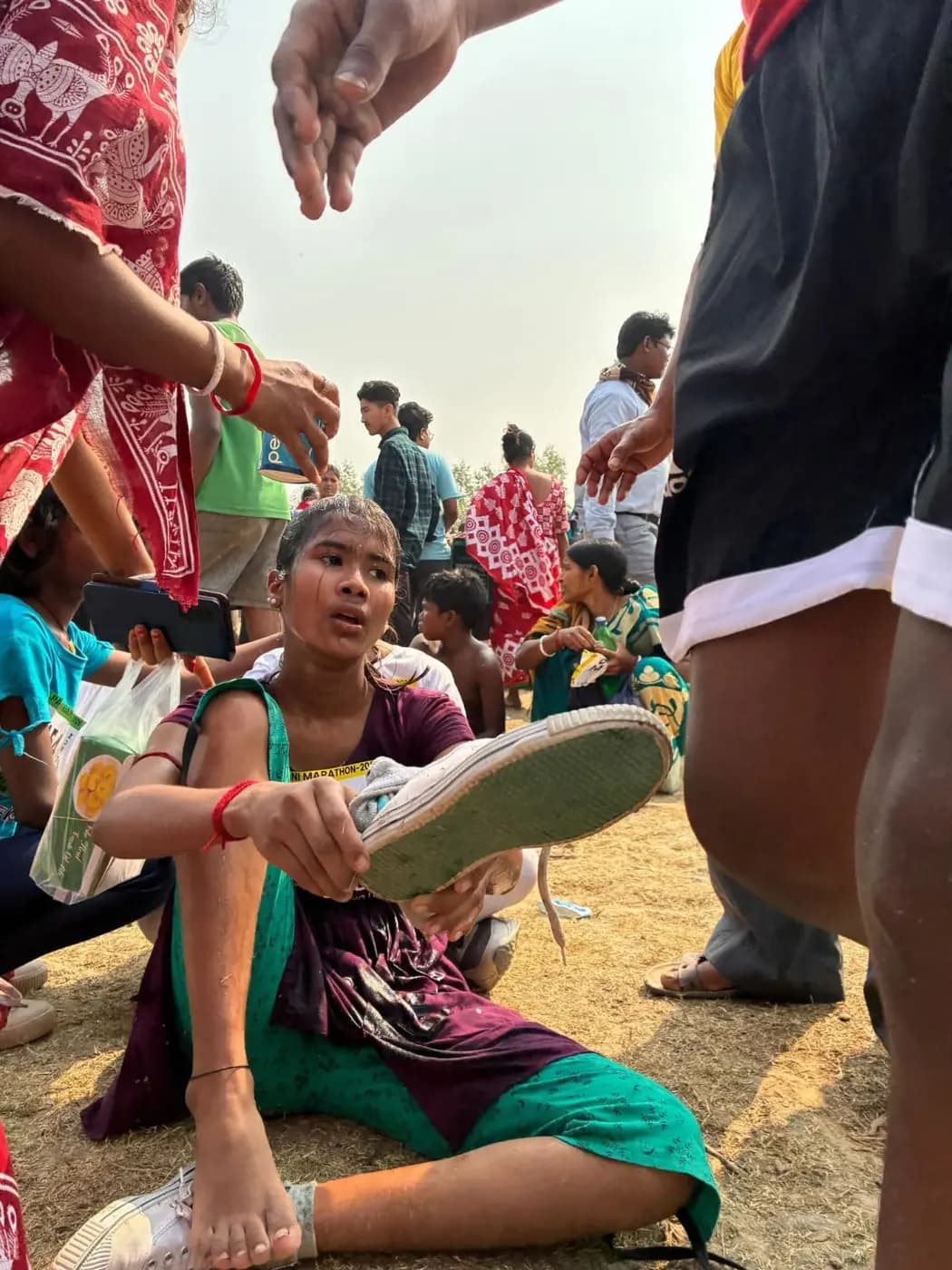 A girl runner, still in her race bib, takes off her canvas shoe on the ground after the run
