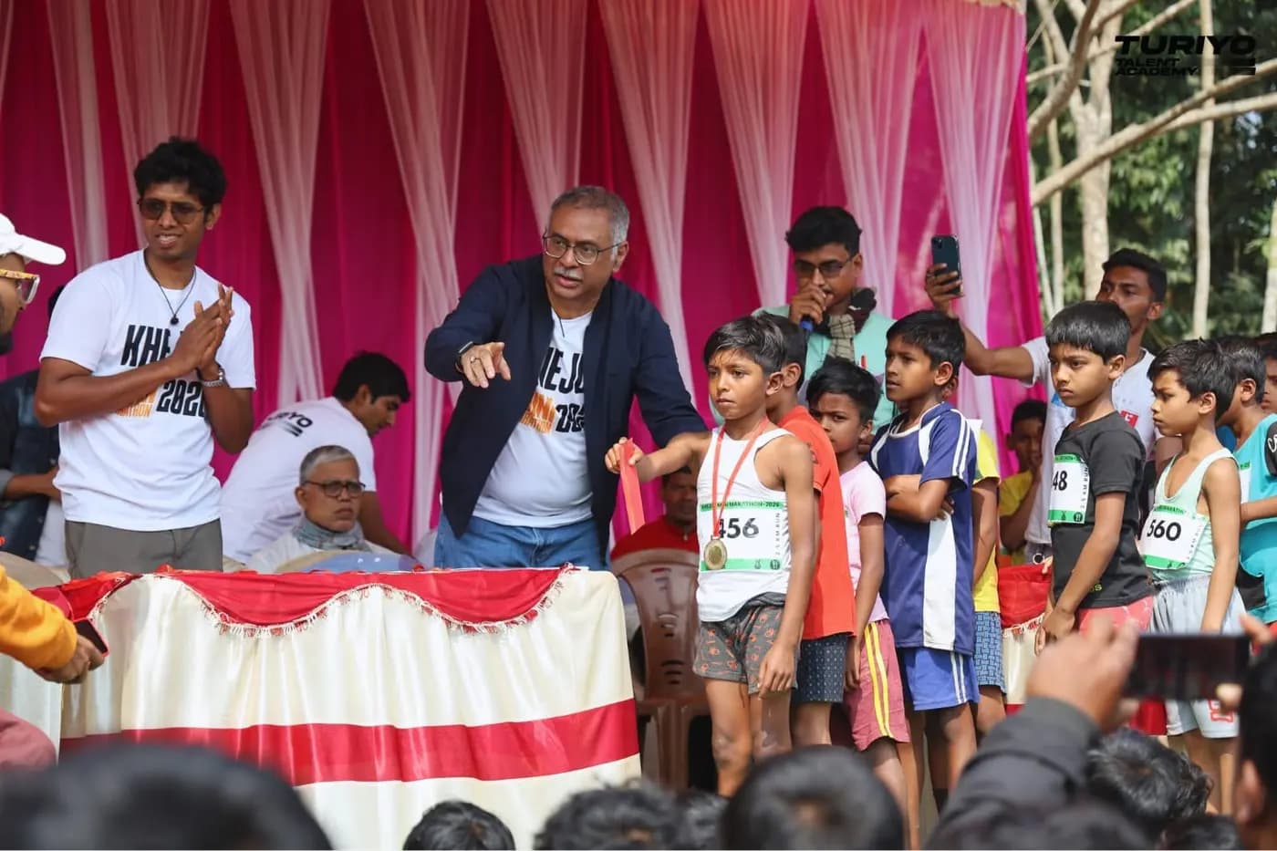 On the prize-giving stage, a boy with his medal stands beside the organisers as other young runners wait their turn