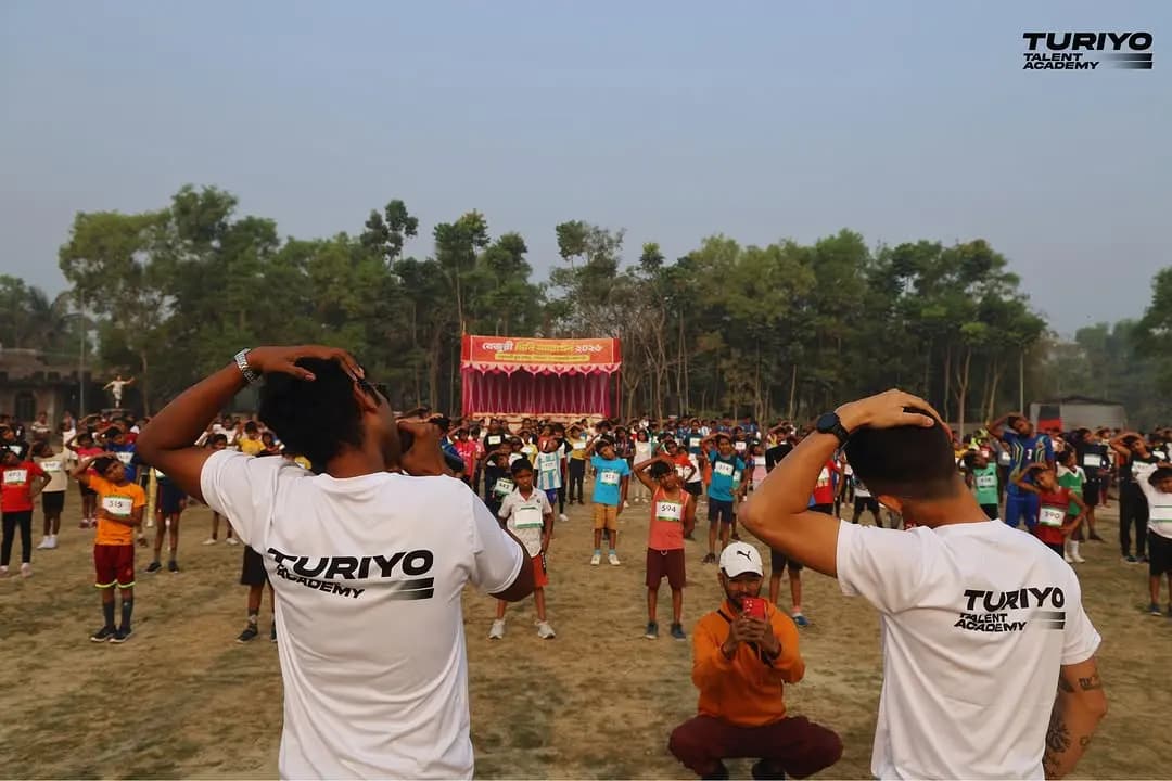 Hundreds of children warm up on the Haludbari High School ground, a volunteer in a TURIYO T-shirt in front