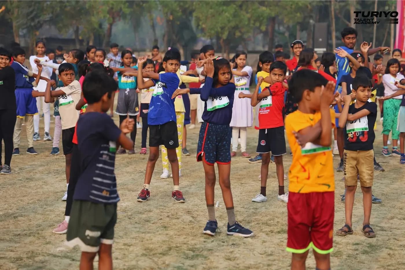 Children in race bibs do arm stretches during the warm-up before the Khejuri Mini Marathon