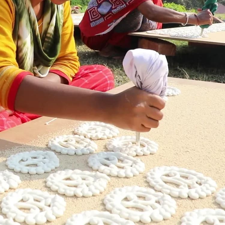 Close-up of a young woman's hand piping Gohona Bori from a cloth bag onto a board dusted with poppy seed, rows of finished patterns in front