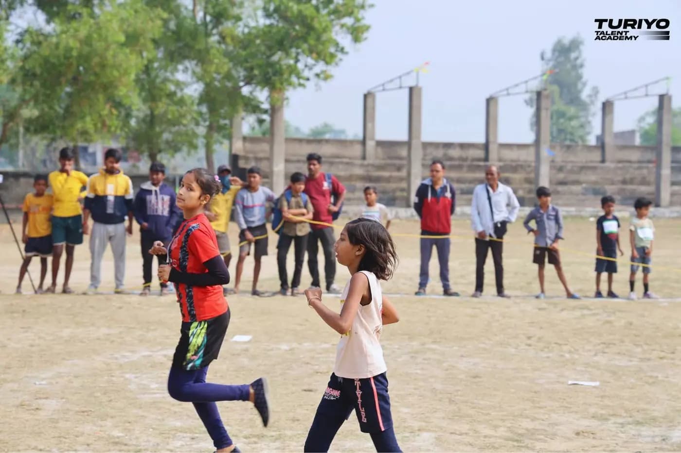 Two girls run across the Haludbari High School ground while boys and parents watch from the rope