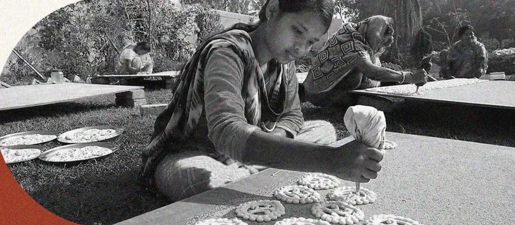 A young woman pipes Gohona Bori patterns onto a board in a village courtyard while other women work behind her